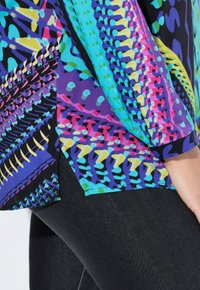 Farverig bluse med abstrakte mønstre i lilla, blå og pink, med en løs pasform og knapdetaljer ved ærmerne, parret med sorte jeans.