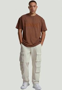 Jovem de pé com as mãos nos bolsos, vestido com uma t-shirt castanha em relevo, calças cargo beje e ténis brancos em frente a um fundo simples.