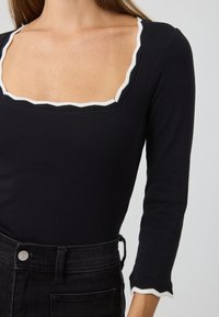Svart bodysuit med en bred, scallop-formet utringning og hvit kant. Lange ermer og en tettsittende design, kombinert med høyt liv svarte jeans.