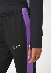 Pantalones deportivos negros con un panel lateral de malla púrpura, que cuentan con una cintura elástica con cordón y un pequeño logo de Nike amarillo en el muslo.