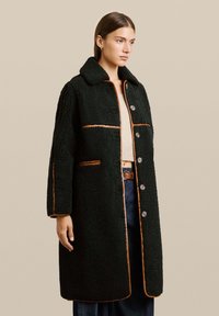 Cappotto nero, strutturato con colletto, caratterizzato da piping arancione e chiusura con bottoni. Il design include due tasche frontali e una vestibilità comoda.