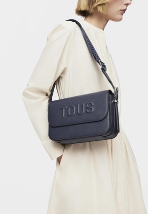 Bolso bandolera de cuero azul marino con el logo "TOUS" en relieve, forma estructurada, cierre de solapa y una correa de contraste. Detalle de superficie texturizada.