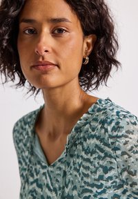 Una donna con i capelli corti e ricci di colore scuro indossa orecchini a cerchio d'argento e una blusa verde a motivo con scollatura ruffles su uno sfondo neutro.