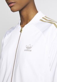 Witte jas met ritssluiting en gouden accenten, voorzien van drie strepen op de schouders, een klein Adidas-logo op de borst en een textuurafwerking van de stof.