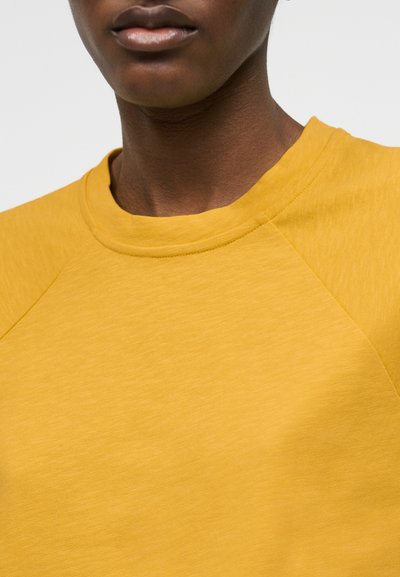 Gros plan sur une personne portant un sweat-shirt à col rond jaune moutarde, montrant l'encolure et la partie supérieure de la poitrine.
