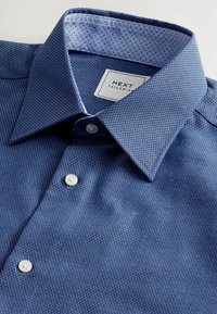 Camicia blu strutturata con motivo a diamante, colletto interno azzurro chiaro, due bottoni bianchi e etichetta "NEXT Tailoring".