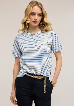 Donna con maglietta a maniche corte a righe blu e bianche con ricamo floreale, pantaloni neri, cintura a catena dorata e orecchini di perle.