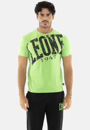 Homme aux cheveux bouclés et foncés portant un T-shirt vert vif avec impression "LEONE 1947" et un pantalon noir avec logo assorti sur fond blanc.