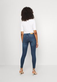 Osoba s kudrnatými vlasy, oblečená v bílém topu, modrých skinny džínách a bílých sandálech na podpatku, stojící čelem k jednoduché bílé zdi.