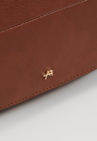 Hnědá kožená taška s hladkým povrchem, ozdobená logem v zlatém tónu. Šití je jemně provedené a precizní.