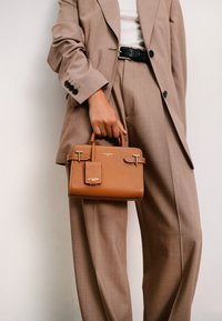 Sac à main en cuir marron avec une forme structurée, doté de quincailleries en argent et d'une étiquette amovible. Porté avec un costume beige.