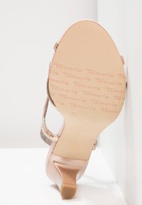 Semelle d'une sandale à talon haut en or rose montrant le logo texturé "Tamaris" et la taille 37, inclinée contre un coin de mur blanc.