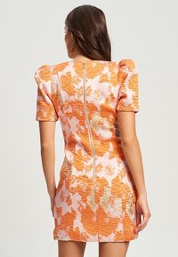 Femme aux longs cheveux bruns portant une robe courte à fleurs texturée orange et rose, avec des manches courtes bouffantes et une fermeture éclair visible dans le dos.