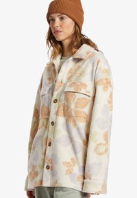 Fleece jacket in cream with pastel floral patterns. Features two chest pockets and brown buttons. Rounded collar, relaxed fit.