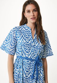 Blå och vit mönstrad klänning med korta ärmar, en krage och ett flätat blått bälte i midjan. Tyget verkar lätt och har en texturerad yta.
