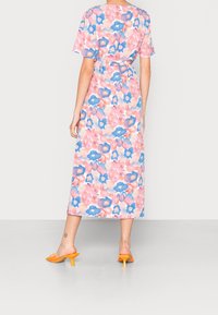 Blumiges Maxikleid in Pink-, Blau- und Pfirsichtönen. Kurze Ärmel, Taillengürtel, fließender Stoff und offene Rückseite. Orangefarbene High-Heel-Sandalen.