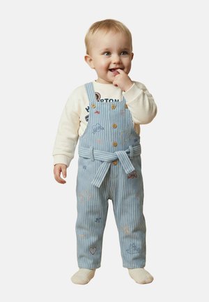 Tout-petit debout, portant une salopette rayée bleue avec de petits motifs brodés et une chemise crème à manches longues, souriant avec le doigt près de la bouche.