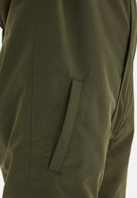 Olijfgroene stoffen broek met een gladde textuur, voorzien van een zijzak die in het ontwerp is geïntegreerd en een strakke, taps toelopende vorm.