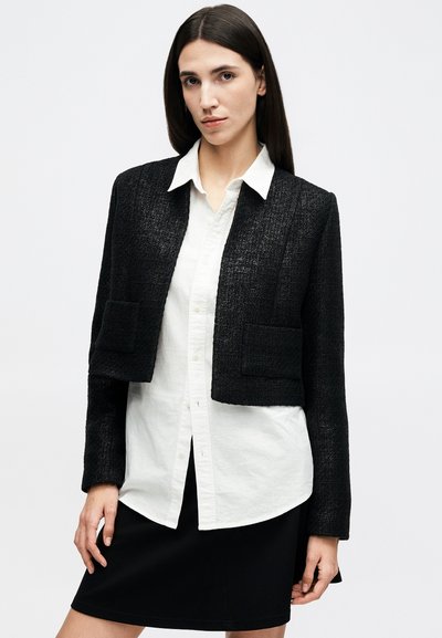 Žena s rovnými tmavými vlasmi má na sebe čiernu textúrovanú krátku bundu, bielu košeľu na zapínanie a čiernu sukňu, stojaca na jednoduchom pozadí.
