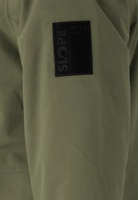Olivgrüner Jackenstoff mit matter Oberfläche. Verfügt über einen schwarzen rechteckigen Aufnäher mit der Prägung "SLOPE" und "STYLE".