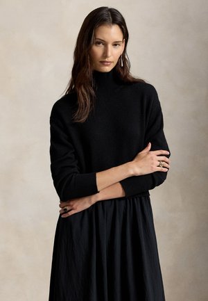Jeune femme aux longs cheveux bruns porte un pull à col roulé noir et une jupe noire, les bras croisés, sur un fond beige uni.