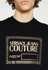 Svart sweatshirt med ribbad halsringning, som har en guld broderad logotyp och kantdesign som inkluderar texten "VERSACE JEANS COUTURE".