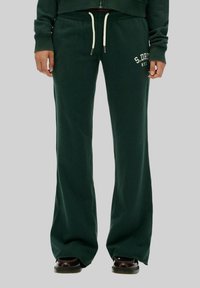 Pantalons de jogging verts avec un design évasé, une taille à cordon et un texte logo blanc sur la jambe gauche. Tissu doux avec une texture lisse.