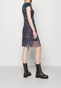 Kvinna som står på sidan iklädd en kortärmad blå blommig klänning och mörkgröna kraftiga Chelsea-stövlar på en ljus bakgrund.