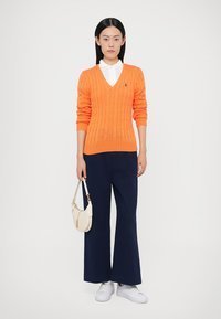 Πλεκτή πορτοκαλί μπλούζα με V λαιμόκοψη και ριμπ μανσέτες, συνδυασμένη με μπλε σκούρο παντελόνι φαρδιάς γραμμής και λευκά αθλητικά παπούτσια. Το μοντέλο κρατά μια μικρή λευκή τσάντα.