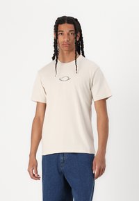 Beige cotton t-shirt with short sleeves and a round neckline, featuring a small oval logo in the center. Worn with blue denim jeans.