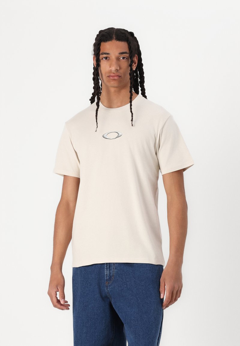 Beige bomull t-skjorte med korte ermer og rund hals, med et lite ovalt logo i midten. Kombinert med blå denim jeans.