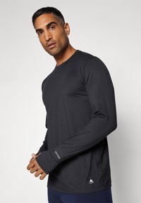 Long-sleeve athletic shirt in solid black, made of synthetic material with a fitted design. Features a logo at the hem and thumbholes in cuffs.