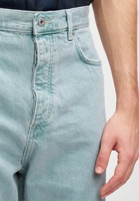 Jeans din denim de culoare bleu deschis, cu talie înaltă și sistem de închidere cu nasturi, având buzunare frontale și cusături vizibile de-a lungul cusăturilor.