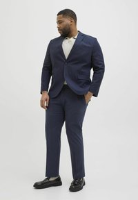 Costume bleu marine avec veste ajustée et pantalon slim; porté par-dessus un polo en tricot beige. Des chaussures noires au fini texturé complètent le look.