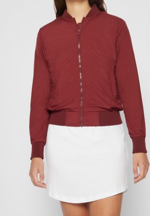 Chaqueta bomber burdeos con cremallera frontal, puños y cintura acanalados. Tejido suave y ligero. Llevada sobre una falda blanca.