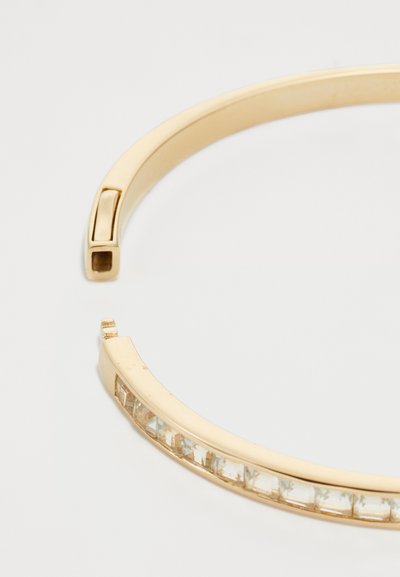 Aranyozott karkötő sima felülettel, négyzet alakú zárómechanizmussal és egy sor átlátszó téglalap alakú kristállyal az egyik szélen.