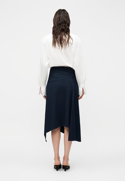 Γυναίκα με μακριά καστανά μαλλιά, ντυμένη με λευκό πουκάμισο με μακριά μανίκια, ασύμμετρη μπλε σκούρα μεσαία φούστα και μαύρα παπούτσια με τακούνι, που στέκεται με την πλάτη προς το μέρος μας.