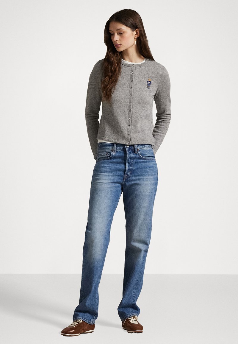 Cardigan grigio con chiusura a bottoni e logo ricamato, indossato sopra una camicia bianca. Abbinato a jeans blu e scarpe marroni. Texture liscia.