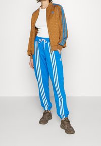 Brun zip-up jacka med ett strukturerat mönster och blåa ränder, kombinerat med blåa sweatpants med vita sidostripes och en justerbar dragsko.