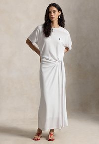 Γυναίκα με μακριά σγουρά μαλλιά που φοράει ένα λευκό φόρεμα κοντομάνικο μέχρι τους αστραγάλους με έναν κόμπο στο πλάι και κόκκινες σανδάλες, που στέκεται μπροστά σε ουδέτερο φόντο.