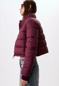 Bordeaux pufferjakke med en cropped design, quiltet tekstur, høj krave og ribbede ærmer. Parret med lyse jeans.