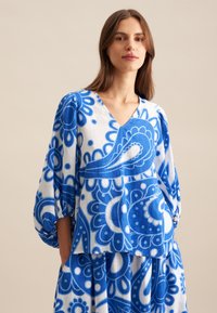 Blusa blu e bianca con motivo, dal colletto a V, maniche a sbuffo e motivi floreali, realizzata in tessuto leggero. Caratterizzata da una vestibilità ampia.