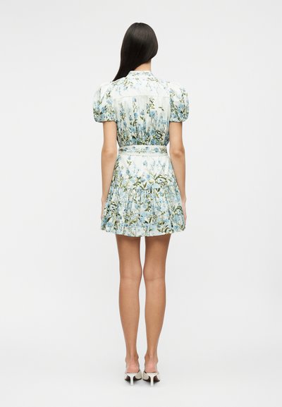 Woman with long dark hair wearing a short, white floral dress with puff sleeves and white heeled shoes, shown from the back.