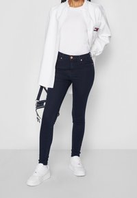 Jeans skinnies azul oscuro de mezclilla, combinados con una camiseta blanca y una camisa blanca abierta. Zapatillas blancas completan el atuendo.