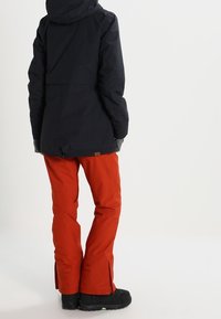 Zwarte waterdichte jas met verstelbare capuchon, gecombineerd met felrode geïsoleerde broek met zijritsen en zwarte laarzen.