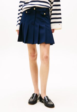 Kvinde iført en mørkeblå plisseret nederdel med guldknapper og sorte loafers, kombineret med en cremefarvet og mørkeblåstribet sweater.