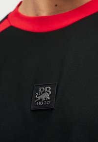 Κοντινό πλάνο μαύρου πουκαμίσου με κόκκινο γιακά, με τετράγωνο μαύρο σήμα που απεικονίζει έναν ταύρο και το κείμενο "PB HUGO" στο στήθος.