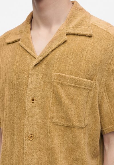 Gros plan d'une personne portant une chemise texturée beige à manches courtes, boutonnée, avec une poche poitrine et un col camp.