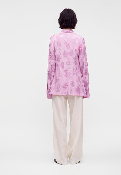 Personne portant un chemisier rose à fleurs à manches longues et un pantalon blanc ample, debout dos à la caméra sur un fond uni.