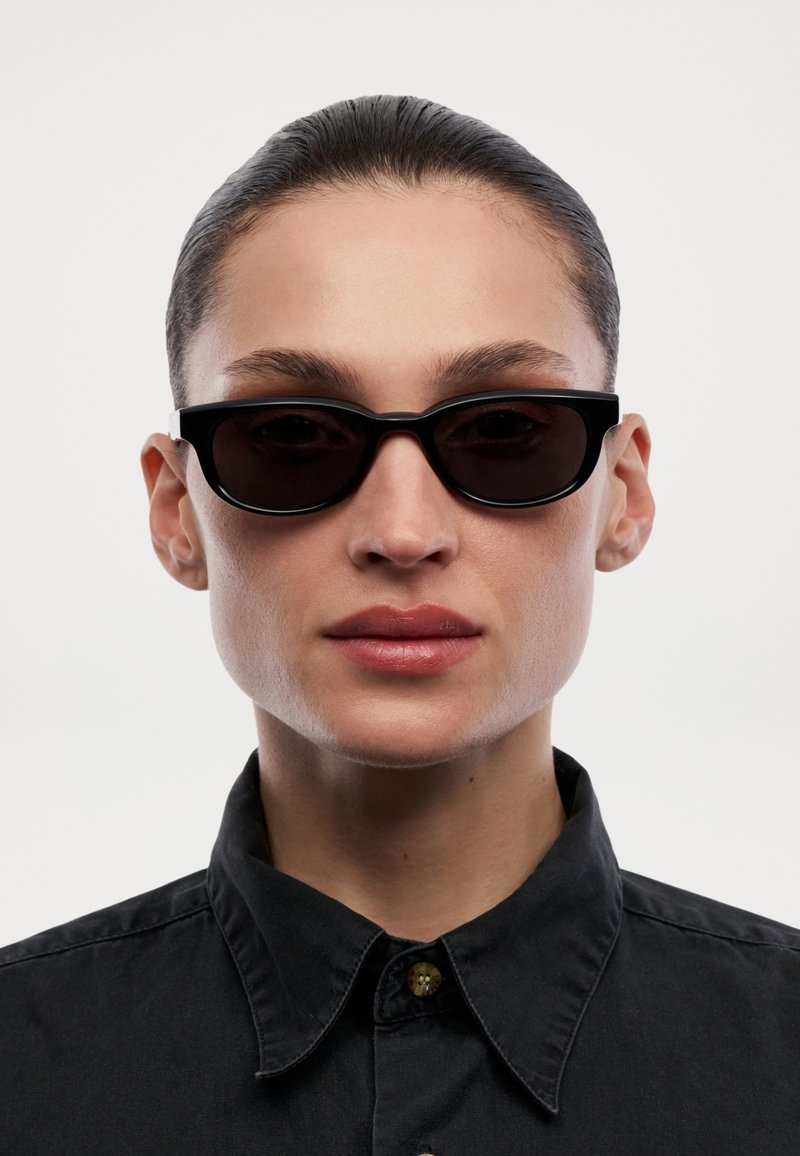 Persona con cabello peinado hacia atrás, usando gafas de sol rectangulares negras y una camisa negra con cuello, frente a un fondo blanco liso.